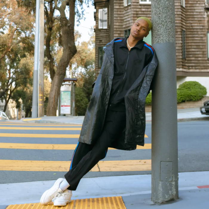 male model in black maison article track suit leaning against pole on street
