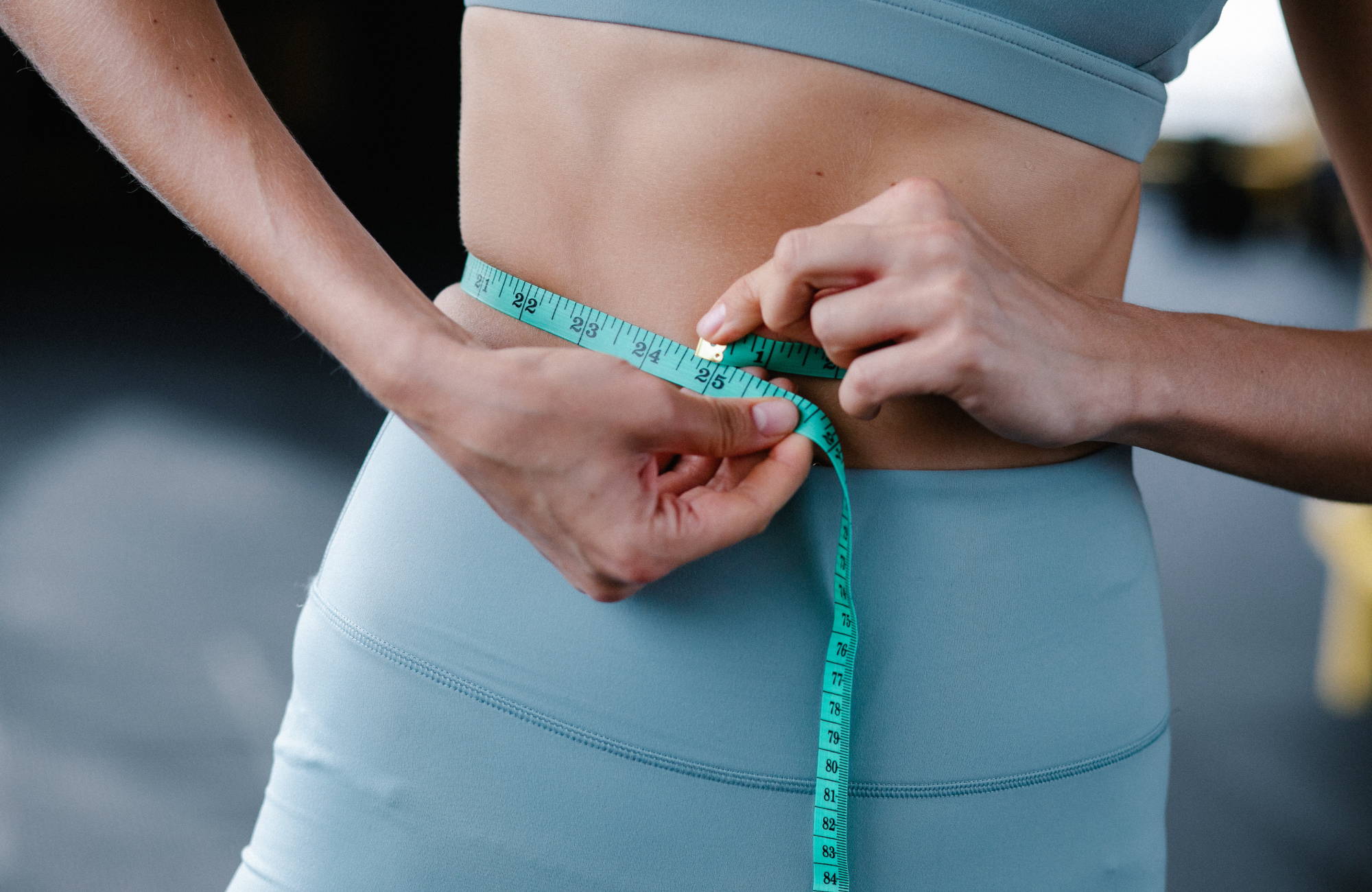 A woman wearing blue yoga pants and a matching sports bra measuring her waist with a teal measuring tape
