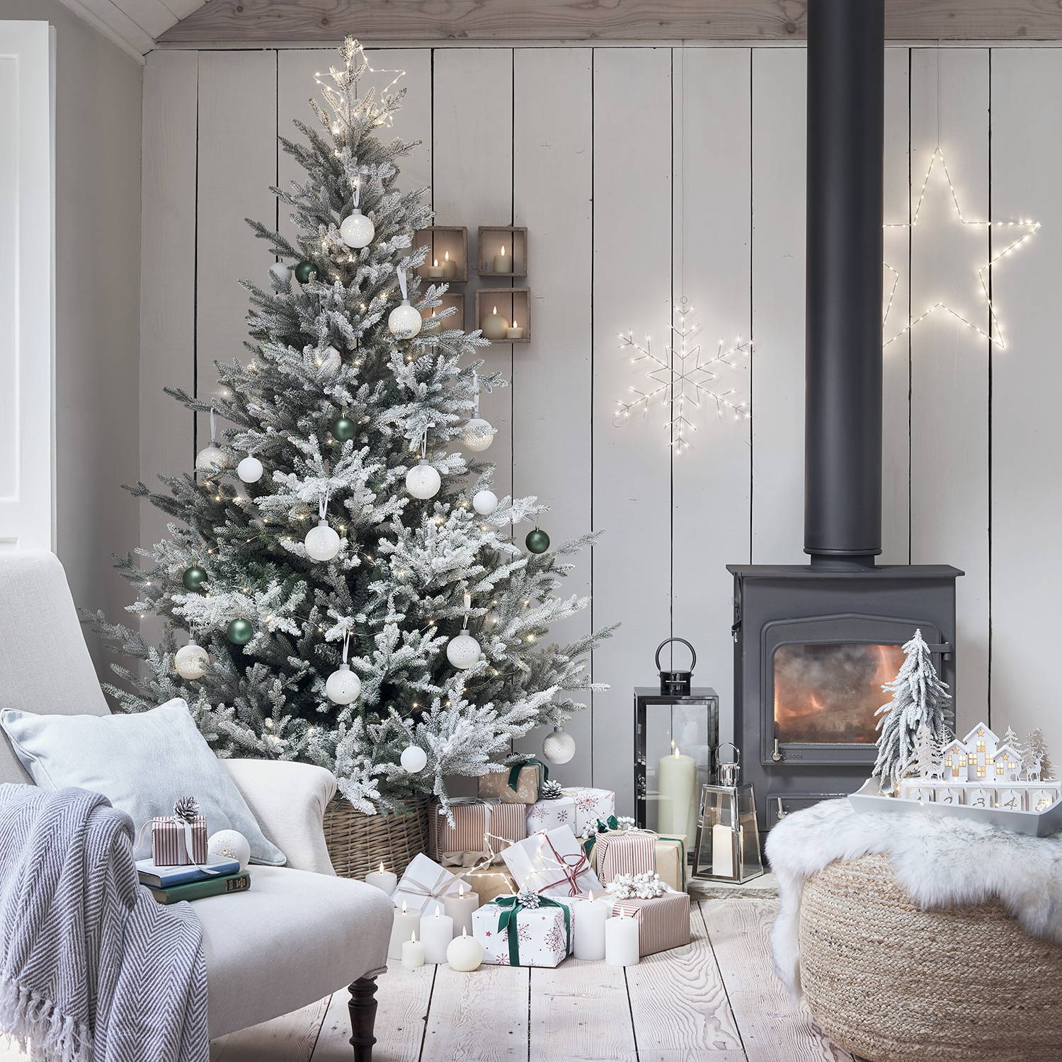 A cosy living room with a frosted christmas tree decorated in neutral whites and silvers with lanterns,  star and snowflake wall lights and a warm log burner