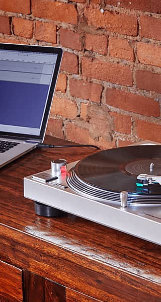 Turntable beside a laptop on a wooden table with a brick wall backdrop.
