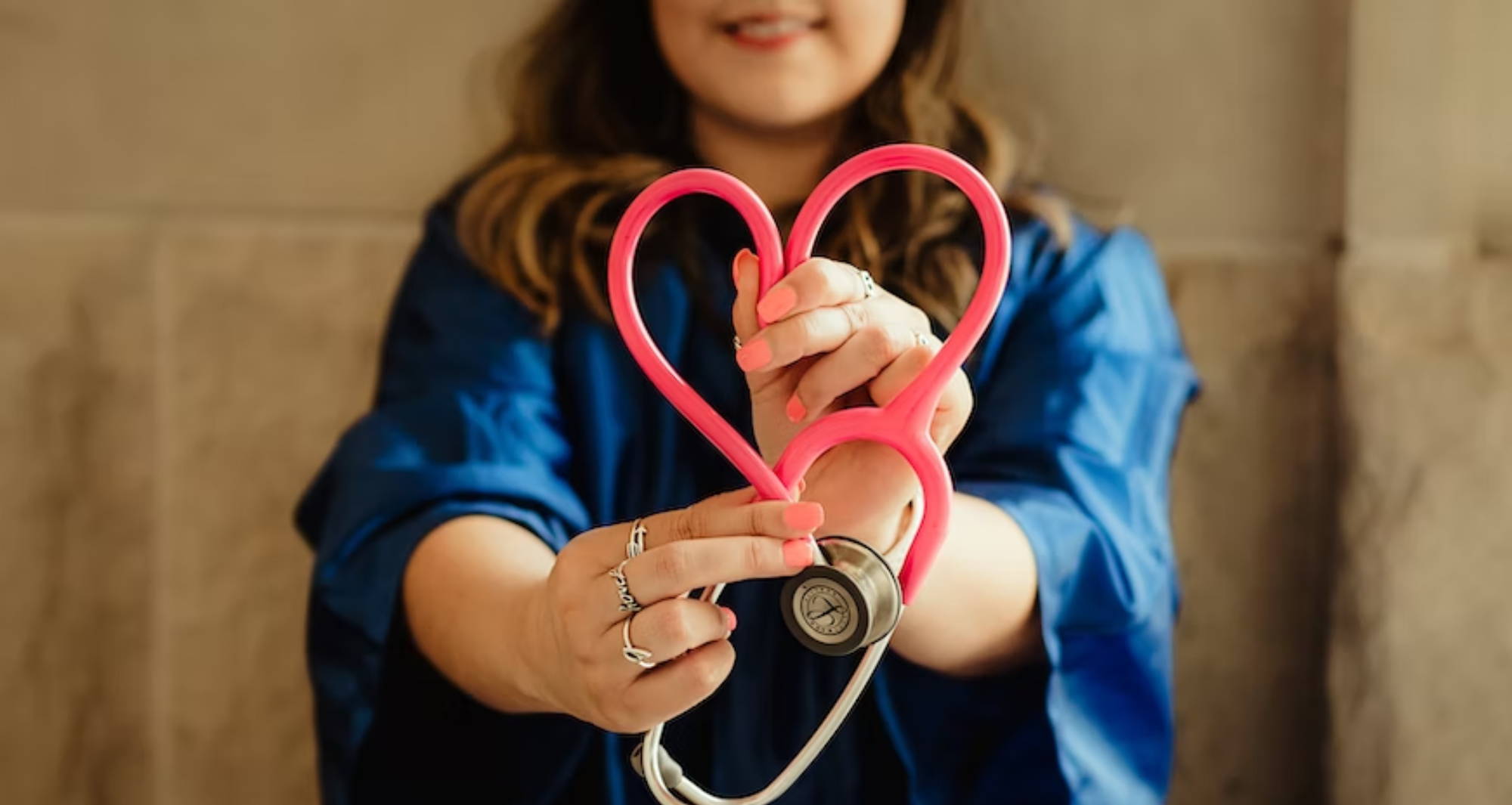 A woman in a blue uniform holds up a pink stethoscope bent into a heart shape.