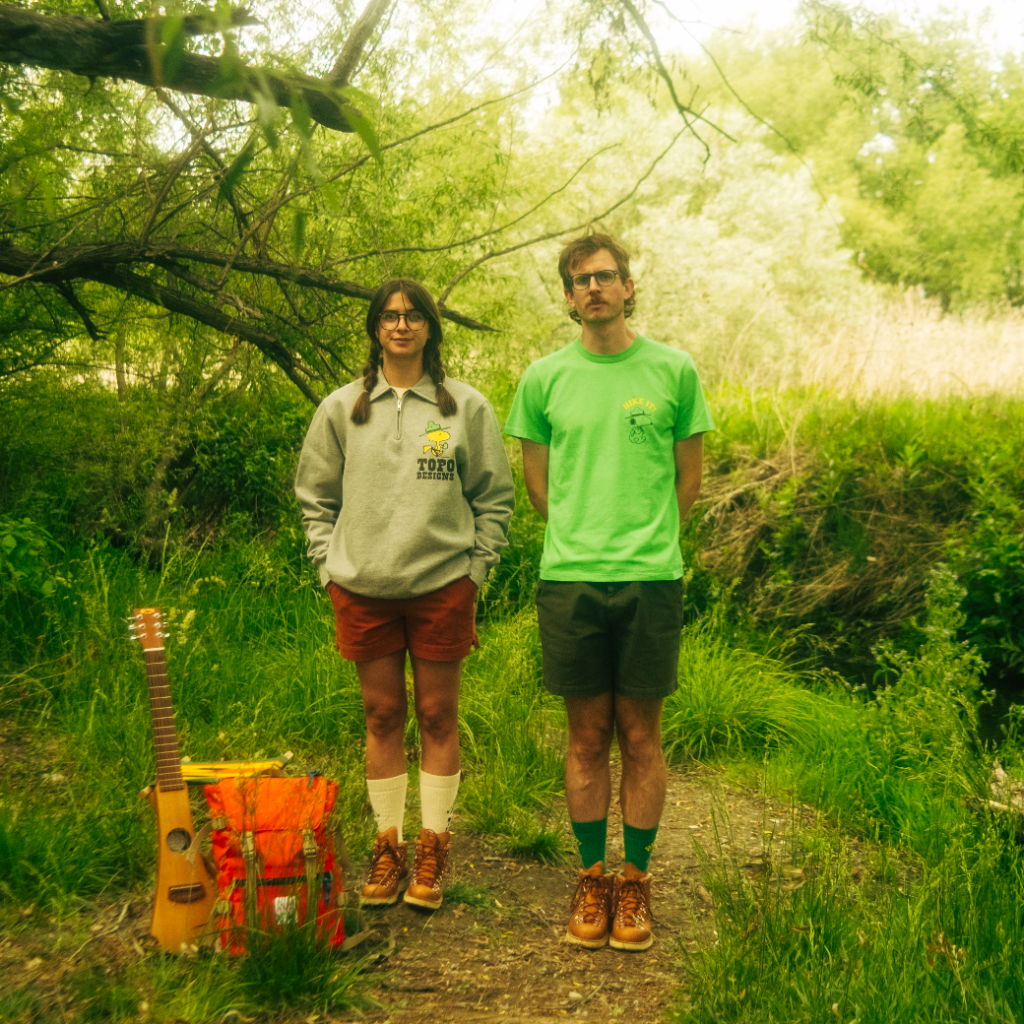 Two people standing in nature, with a guitar and a bucket nearby.