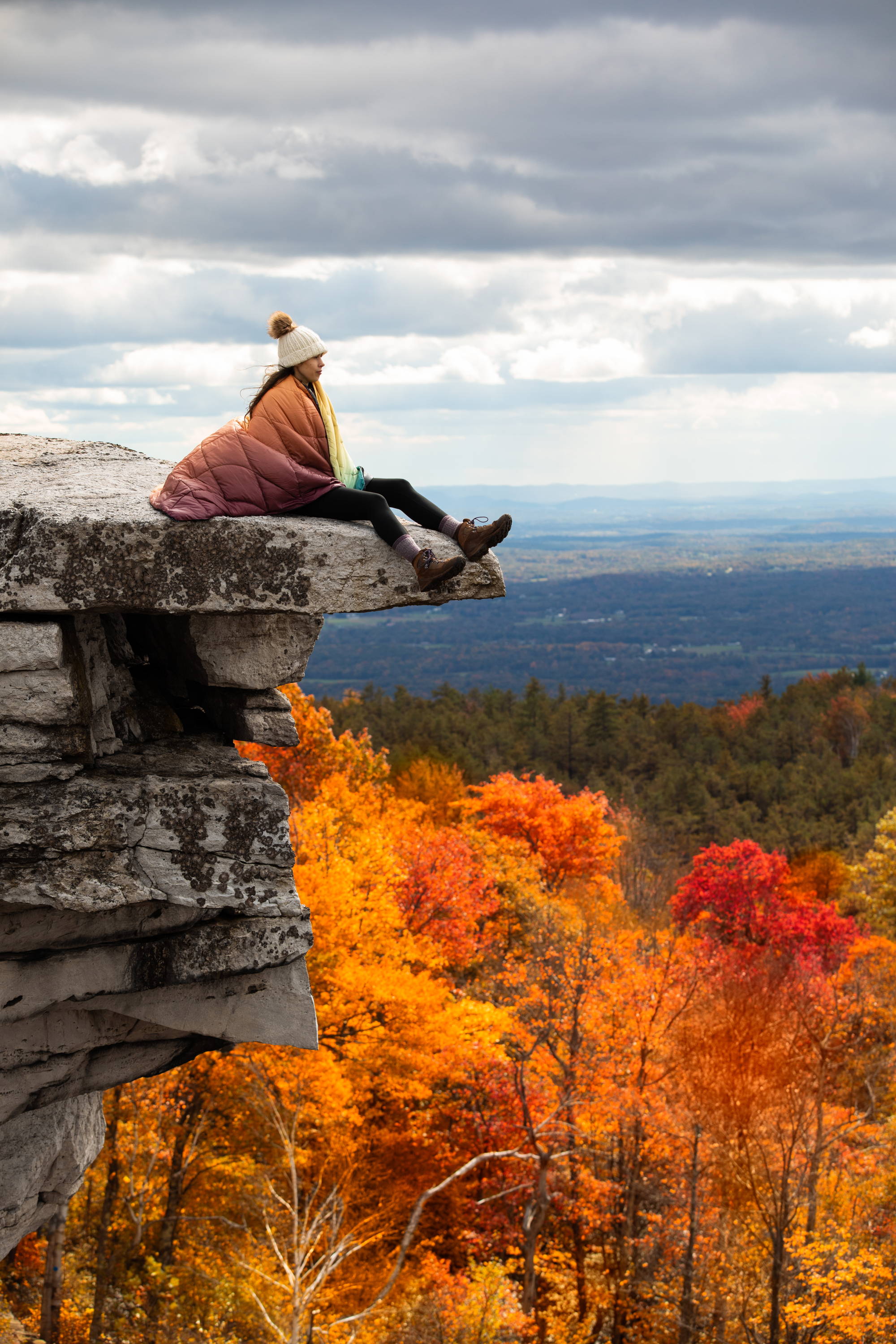 Woman wearing Rumpl blanket while sitting on cliff overlooking autumn landscape