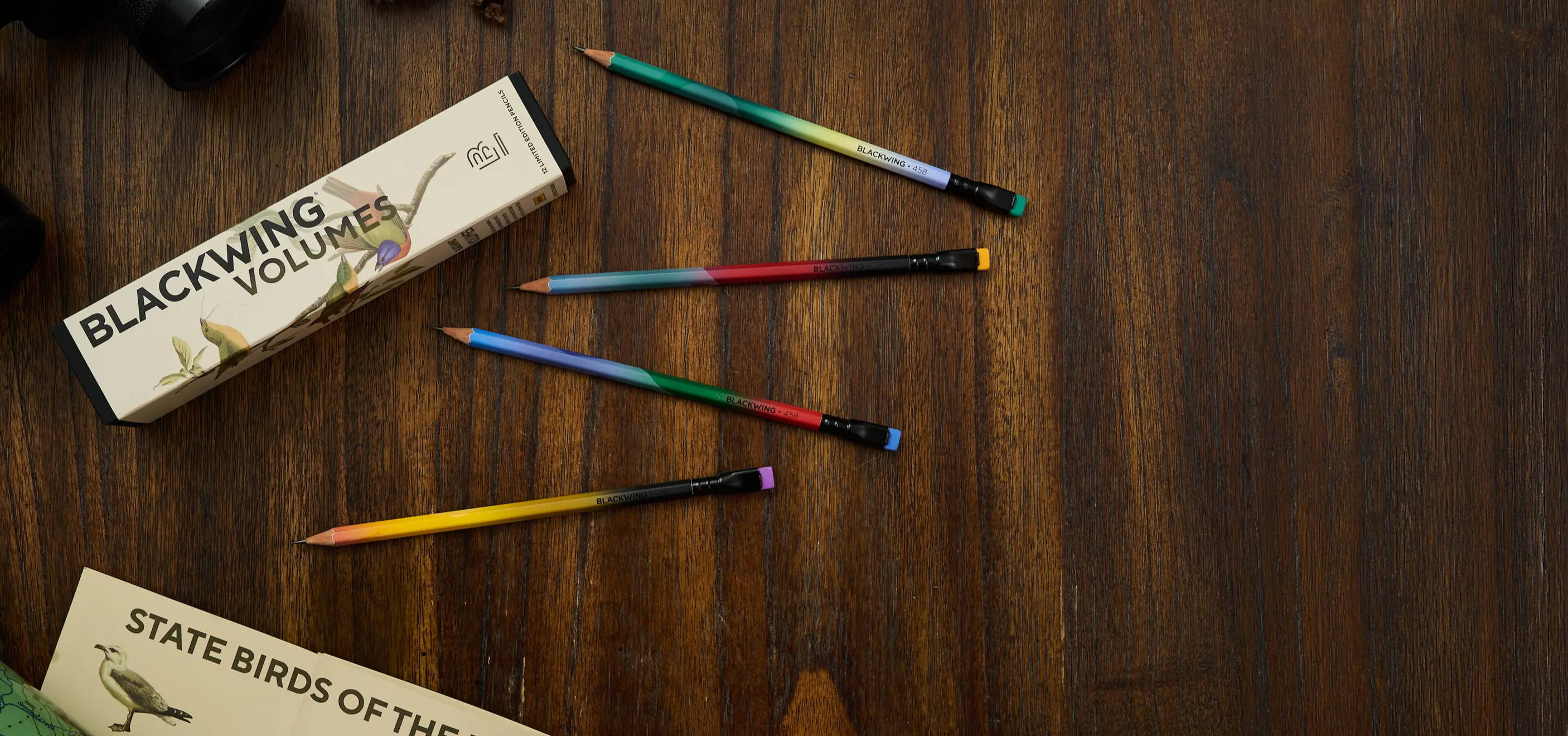 Blackwing Vol. 458 packaging and 4 gradient design pencils laid out on a wood desk. Binoculars, state bird chart, and map arranged above and below the centerpiece set.