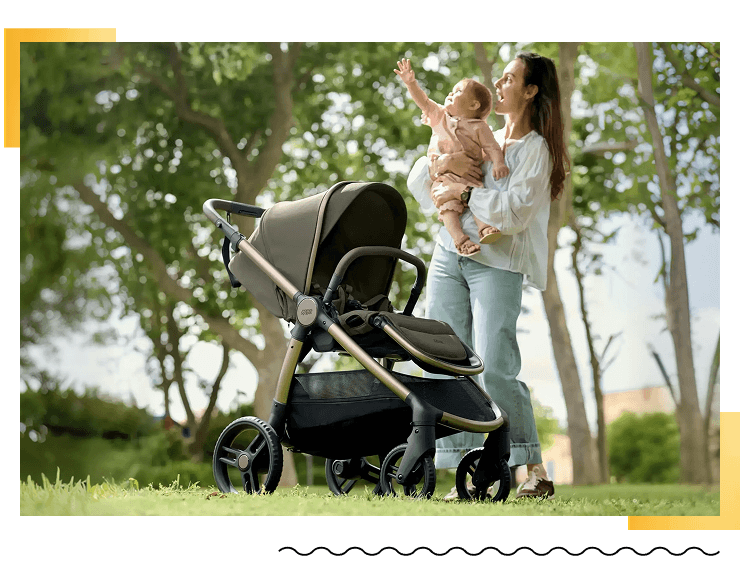 A mother holding a baby stands beside a stroller in a park.