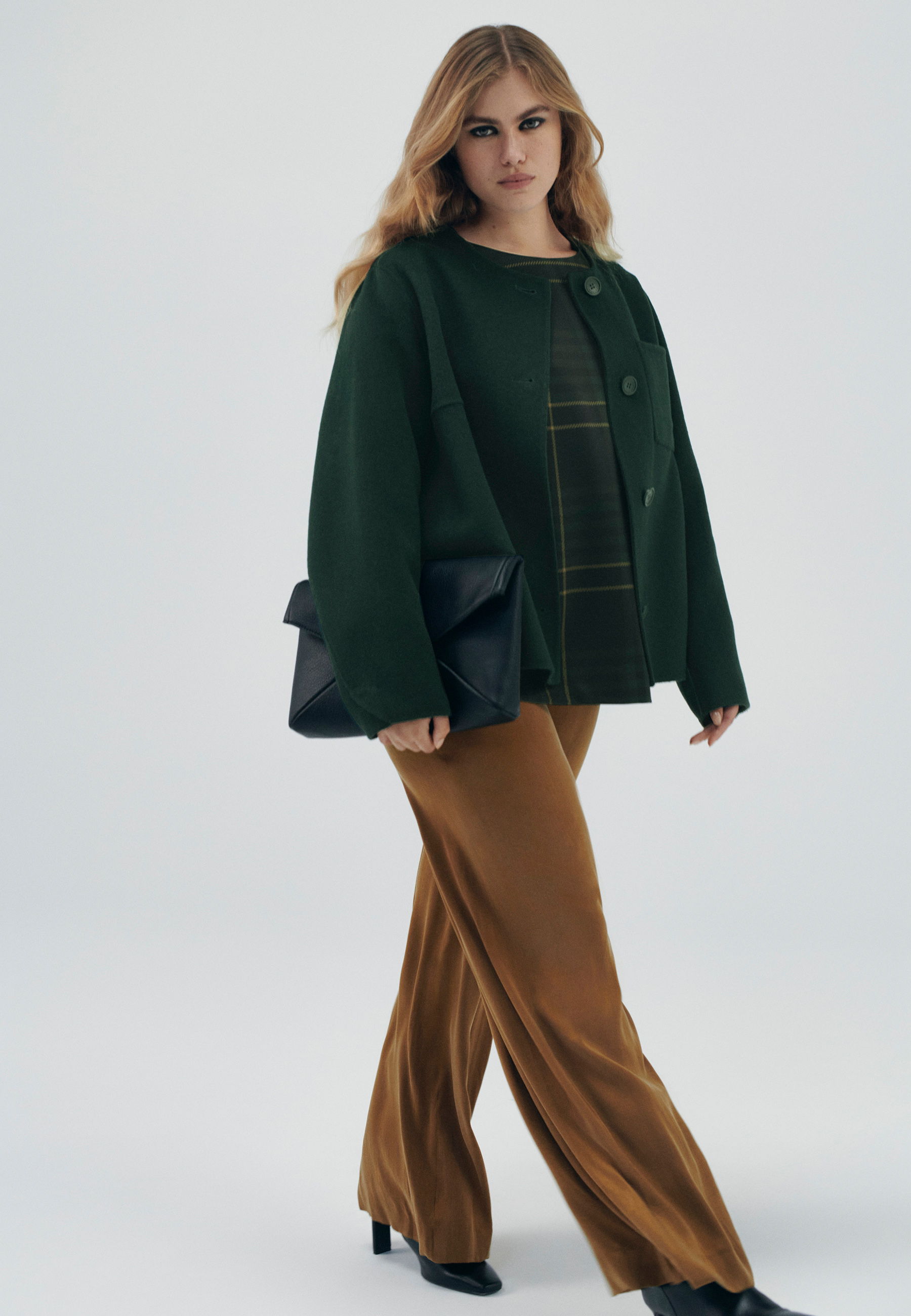 Woman in green jacket and brown pants, holding a black handbag.
