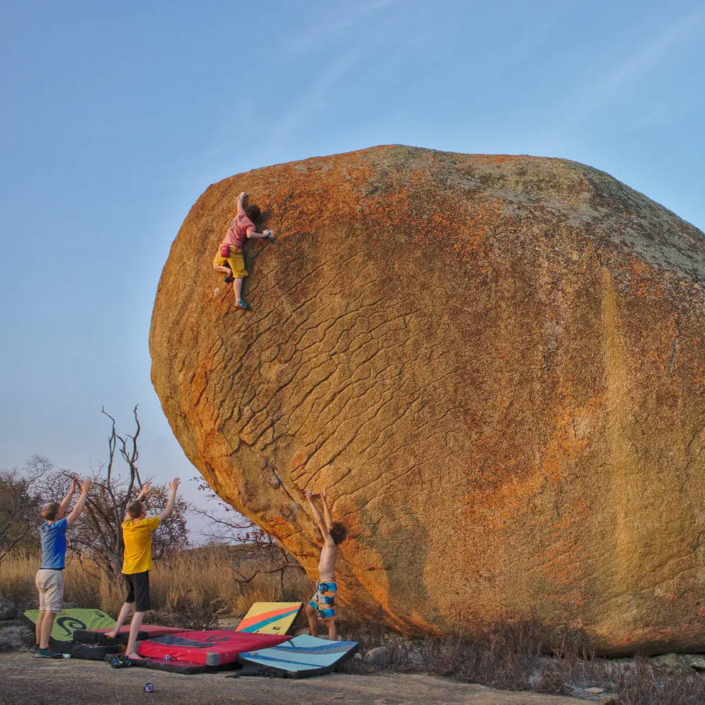 Climber is at the top of a highball boulder with 3 spotters lifting their arms to try and protect a fall