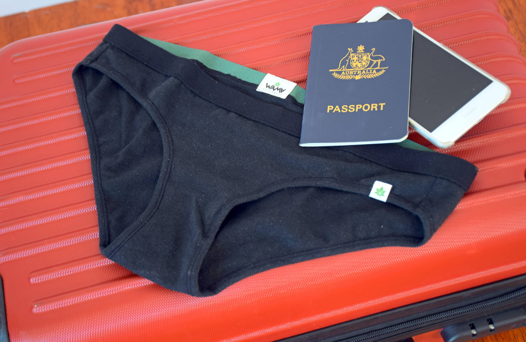  A pair of black underwear, an australian passport, and a white phone laid out on top of a red suitcase